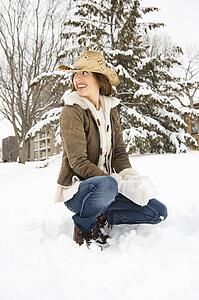 Caucasian young adult female looking over shoulder while kneeling in snow with snowball and wearing straw cowboy hat.