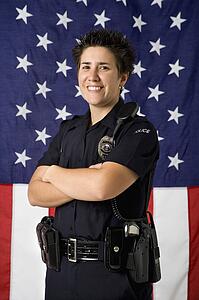 Portrait of mid adult Caucasian policewoman standing with arms crossed with American flag as backdrop smiling at viewer.