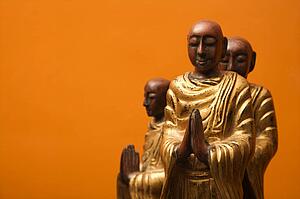 Three wooden statues of Buddhist disciples against orange wall.