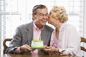 Mature Caucasian woman kissing mature Caucasian man while holding birthday cake.