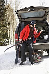 Caucasian male teenager sitting on back of SUV holding skis.