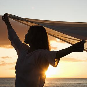 Attractive Caucasian mid-adult woman holding up fabric in breeze silhouetted by sunset beside ocean.
