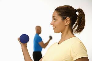 Close up of smiling mid adult woman exercising using dumbbells with mid adult man in background.