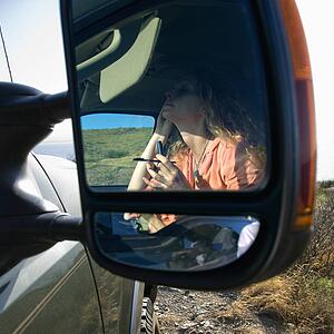 Reflection in vehicle side mirror of Caucasian woman applying mascara.