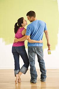 Attractive couple standing in front of partially painted wall with arms around each other smiling