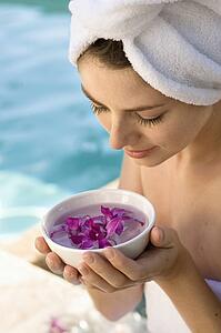 Woman wearing towel around head and body holding bowl of purple orchids next to pool