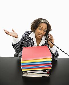 Businesswoman sitting at office desk with large stack of work talking on telephone and gesturing.