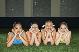 Four multiethnic girls lying on ground in indoor gym with heads on hands smiling.