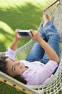 Smiling pretty young adult Caucasian brunette female lying in hammock looking up at veiwer and using PDA.