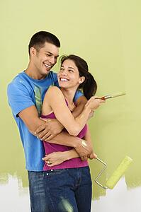 Couple smiling and embracing in front of partially painted interior wall holding paintbrush and roller.