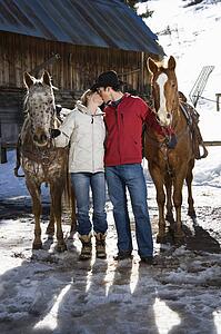 Couple kissing and holding horses with stable in background.