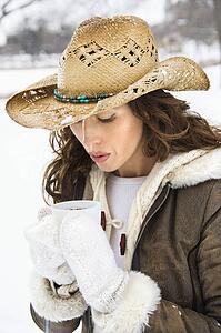 Female outdoors in snow blowing into coffee cup and wearing straw cowboy hat