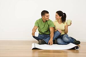 Attractive couple sitting on home floor with coffee cups looking at house plans and smiling at each other.