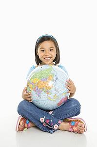 Asian girl sitting on floor holding Earth globe in her lap.