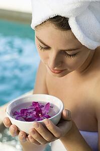 Caucasian mid-adult woman wearing towel around head and body holding bowl of purple orchids next to pool.