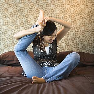 Woman sitting on bed trying to place foot behind her head