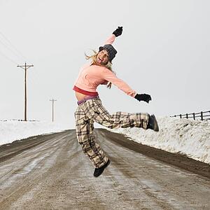 Young woman in winter clothes standing on muddy dirt road jumping and smiling.