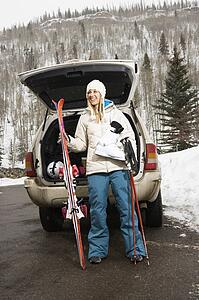 Woman in winter clothes standing holding ski equipment and smiling by automobile
