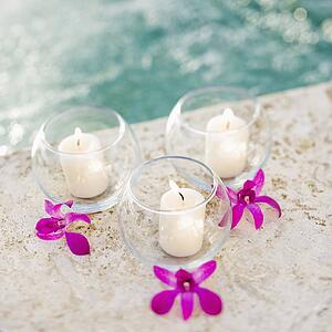 Three lit candles in glass bowls with purple orchids next to pool.