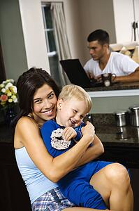 Woman hugging toddler son with father on laptop in background