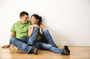 Attractive young adult couple sitting close on hardwood floor in home smiling