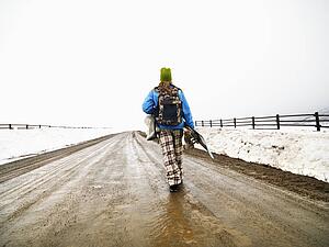 Young woman in winter clothes walking down muddy dirt road holding snowboard and boots.