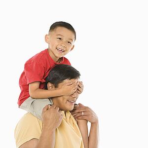 Asian boy sitting on father's shoulders with hands over his eyes in front of white background.