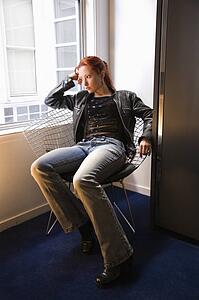 Pretty redhead young woman sitting indoors on metal chair by window looking out