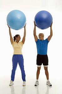 Mid adult man and woman holding blue exercise balls over their heads looking at each other smiling
