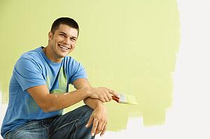 Smiling man kneeling in front of partially painted wall holding paintbrush