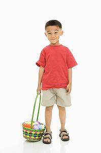 Asian boy standing against white background holding Easter basket.