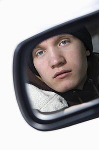 Caucasian male teenager looking at himself in side view mirror of car.