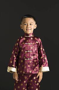 Asian boy in traditional attire standing against black background.