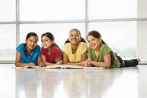 Preteen girls of mutiple ethnicities sitting together on floor with schoolwork smiling at viewer.