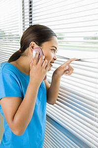 Side view of girl looking through blinds out window while talking on cell phone