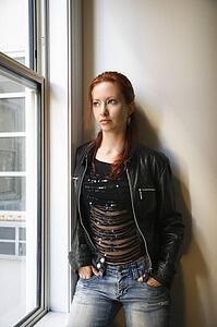 Pretty redhead young woman standing indoors by window looking out leaning against wall with hands in jean pockets.