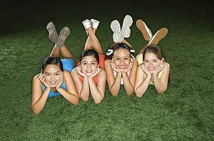 Four multiethnic girls lying on ground in indoor gym with heads on hands smiling.