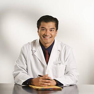 Asian American male doctor sitting at desk with charts smiling and looking at viewer.