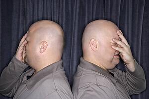 Profile of bald identical twin men standing back to back and grimacing with hands to head.