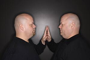 Bald mid adult identical twin men standing face to face with hands touching.