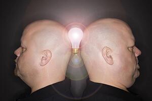 Mid adult identical twin men standing back to back and balancing a light bulb between them.