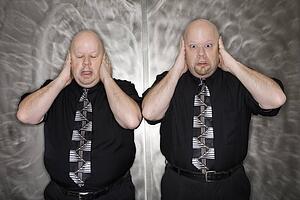 Bald mid adult identical twin men standing with hands covering ears making facial expression.