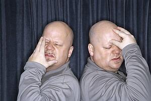 Bald identical twin men standing back to back and grimacing with hands to head