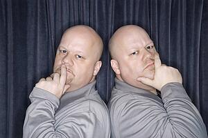 Bald identical twin men standing back to back and looking at viewer with hands to mouth.
