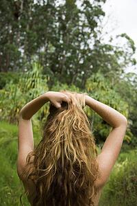 Back view of woman in lush forest holding up long wavy hair.