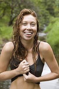 Wet Caucasian young adult woman standing in stream wringing water out of her shirt smiling and looking at viewer.