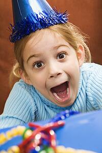 Caucasian girl wearing party hat holding gift and looking at viewer with excited expression.
