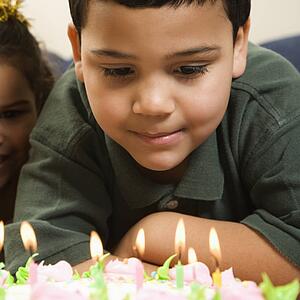 Boy looking down wishfully at lit candles of birthday cake with girl peeking in over his shoulder