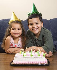 Hispanic girl and boy wearing party hats sitting in front of birthday cake smiling and looking at viewer.