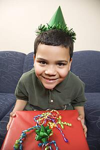 Boy wearing party hat holding large birthday present smiling and looking at viewer